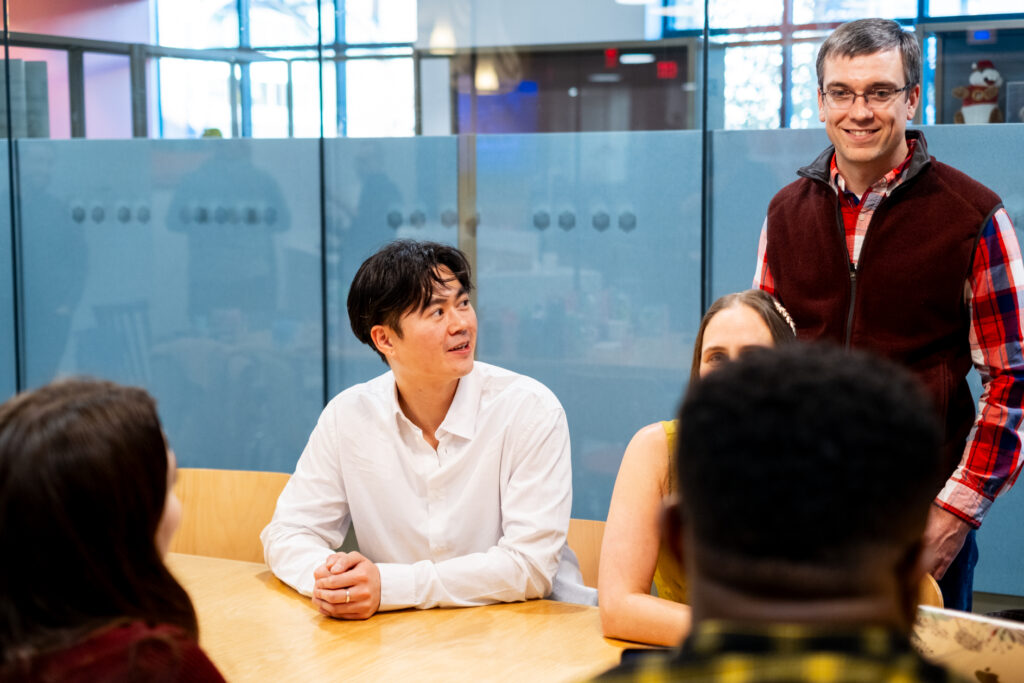 One person sitting at a table with another person beside him standing