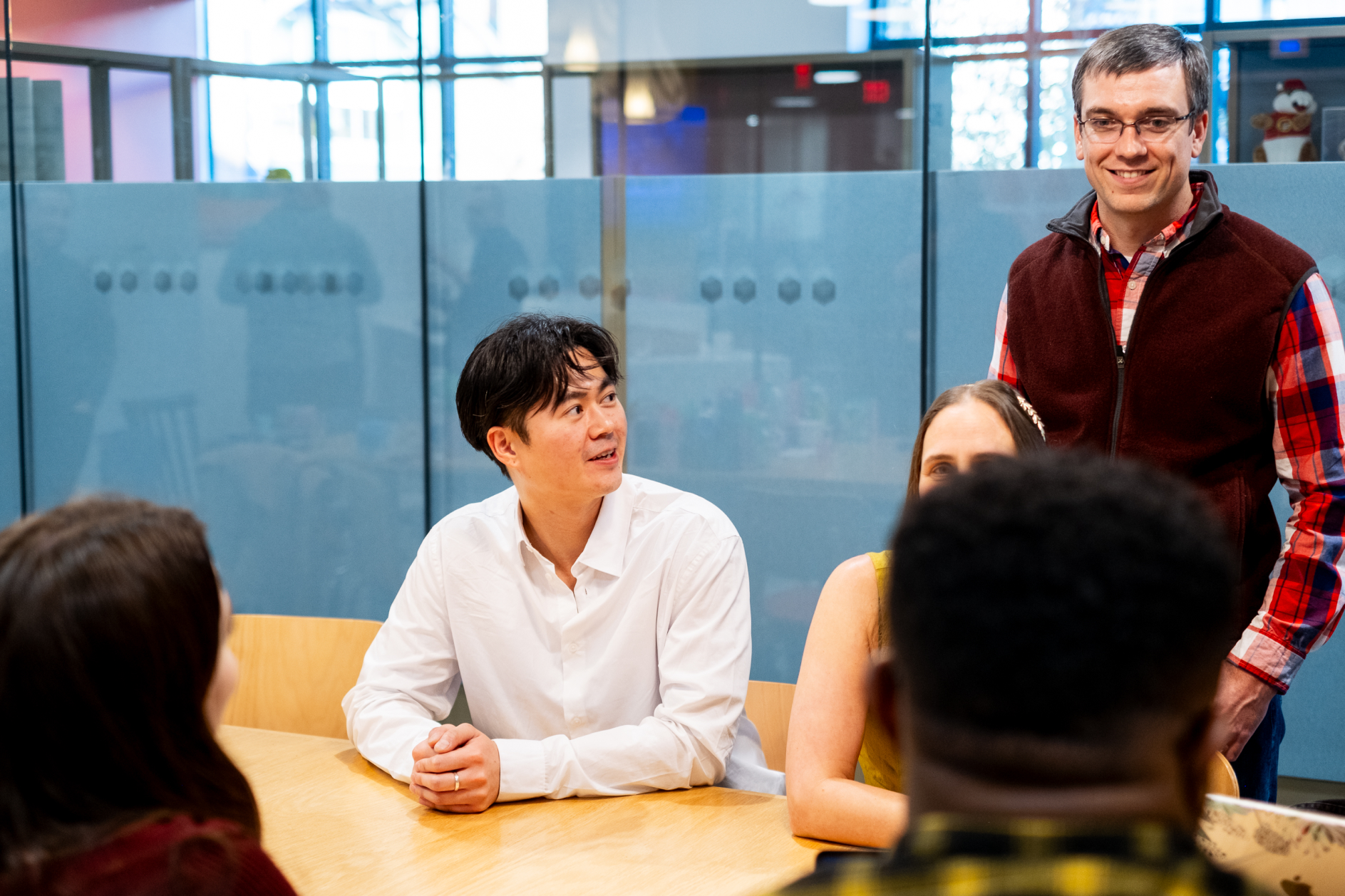 One person sitting at a table with another person beside him standing